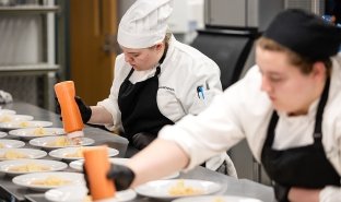 Two Culinary students dressed in chefs' uniforms and hats plate food at an event at Seasoned.