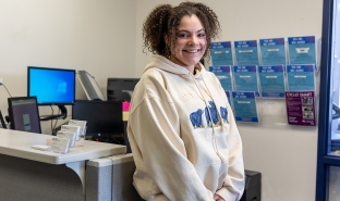 Sophia Goldsmith smiles at the camera in the Student Activities office. She has dark curly hair and wears a hooded sweatshirt.