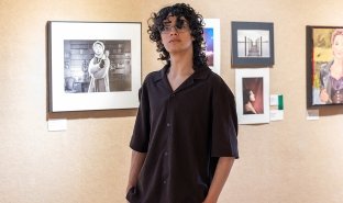 Ivan Aviles stands in front of some of his photography in the 2026 Annual Juried Student Art Exhibition in the Visual Arts Gallery, with his hands in his pockets.
