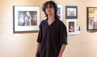 Ivan Aviles stands in front of some of his photography in the 2026 Annual Juried Student Art Exhibition in the Visual Arts Gallery, with his hands in his pockets.