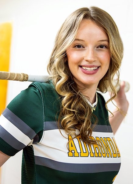 A woman with long, wavy brown hair is smiling at the camera, with a softball bat over her shoulders