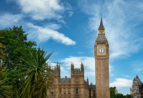 Big Ben is seen in London, with trees framing the left side of the image and blue skies with wispy clouds