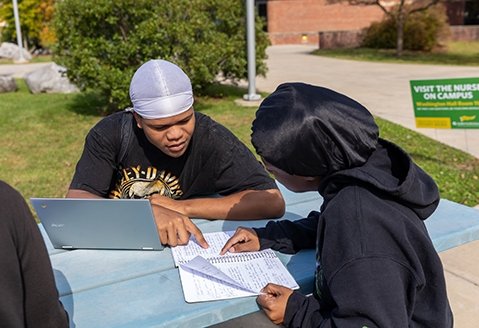 Two students review notes at a picnic table on campus
